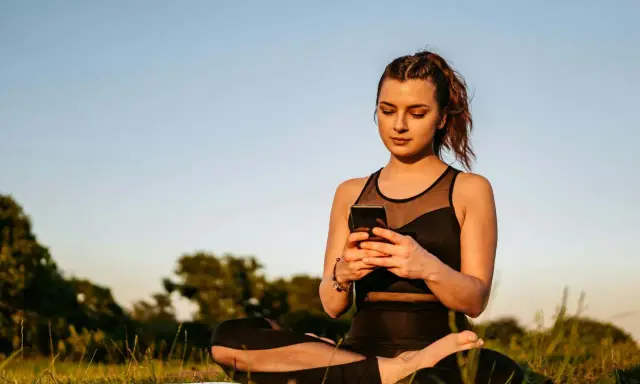 Kvinna i lotusställning på en matta utomhus, med en mobiltelefon i händerna. En modern typ av meditation.