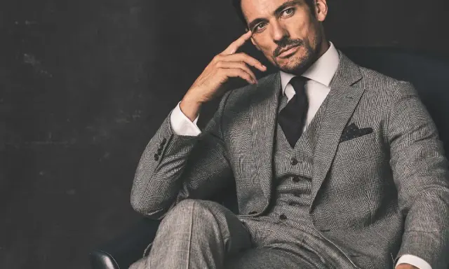 A man in a grey suit, black shirt, and tie, with a thoughtful expression, sits in a chair.