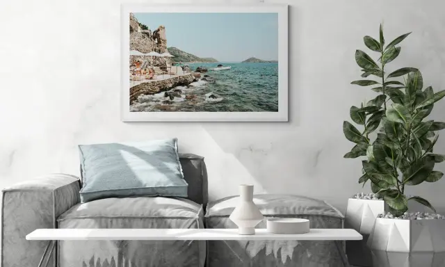 A framed print of a coastal scene, reminiscent of fine art photography galleries, hangs above a sofa. The artwork depicts a rocky shoreline with people relaxing under umbrellas.