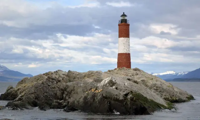 Faro a strisce rosse e bianche su un'isola rocciosa nel Canale di Beagle, con montagne innevate sullo sfondo.