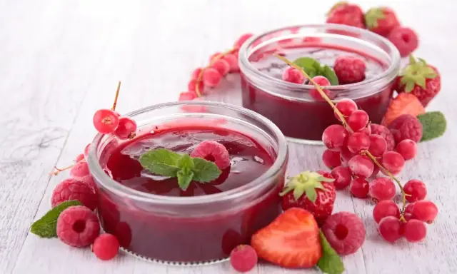 Due coppette di coulis di lamponi, guarnite con lamponi freschi, ribes e foglie di menta, circondate da fragole e frutti di bosco.