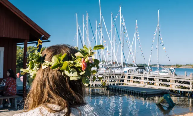 En flicka med blomsterkrans ser ut över en hamn med segelbåtar. En perfekt dag för en resa till Åland.