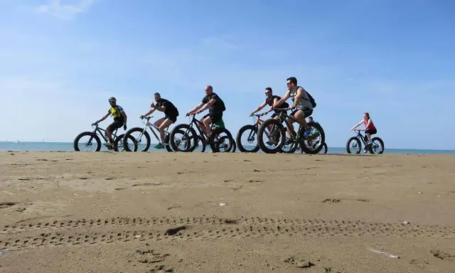 Gruppo di ciclisti su fat bike lungo la ciclabile di Caorle, sulla sabbia dorata con il mare sullo sfondo.