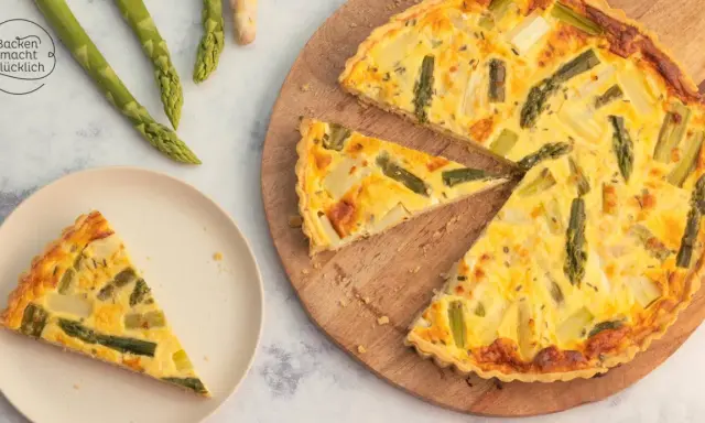 Herzhafte Spargel Quiche mit frischem Spargel, goldbraun gebacken. Ein Stück auf einem Teller, der Rest auf einem Holzbrett.