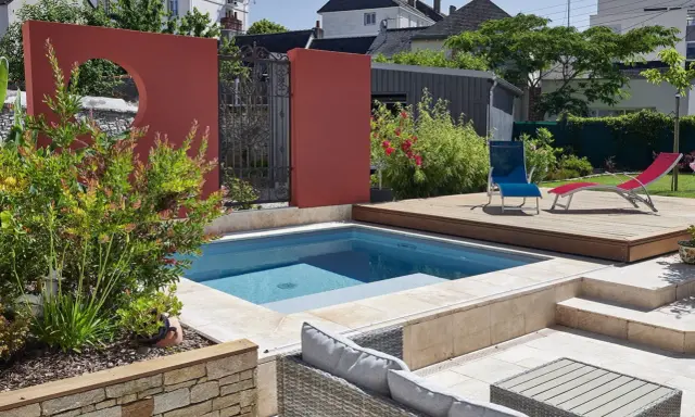 Une piscine moderne avec une belle décoration autour piscine : mur rouge, terrasse en bois, mobilier de jardin et végétation luxuriante.