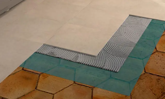 Dettaglio di un pavimento in fase di posa: nuove piastrelle beige sopra uno strato di colla, membrana impermeabilizzante turchese e vecchie piastrelle esagonali color terra.