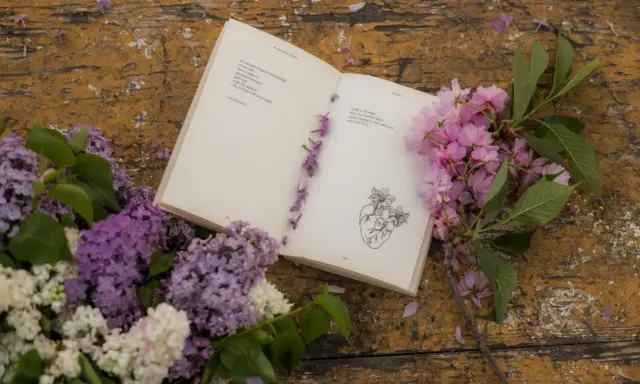 Un libro aperto con poesie d'amore passionali anonime, decorato con fiori lilla e rosa su un tavolo di legno consumato.