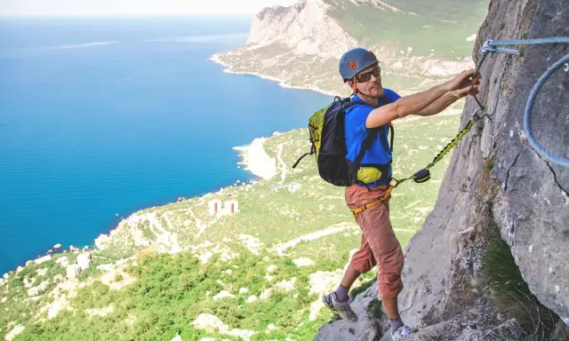 Mężczyzna na via ferrata Krucze Skały, zmagający się ze stromą ścianą skalną, z widokiem na błękitne morze i zielone wybrzeże.