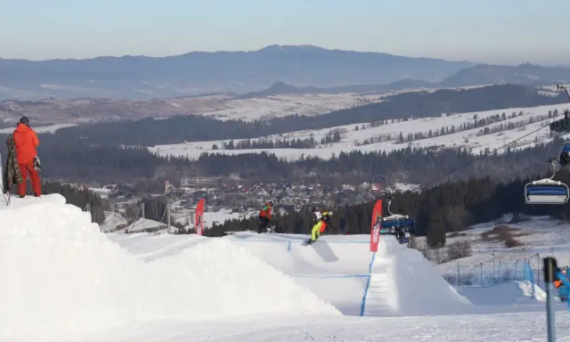 Snowboardziści na stoku z wyciągiem krzesełkowym i widokiem na góry i miasteczko.