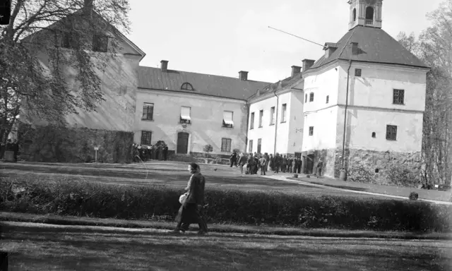 En grupp människor samlas framför Rydboholms slott. En kvinna går över gräsmattan.