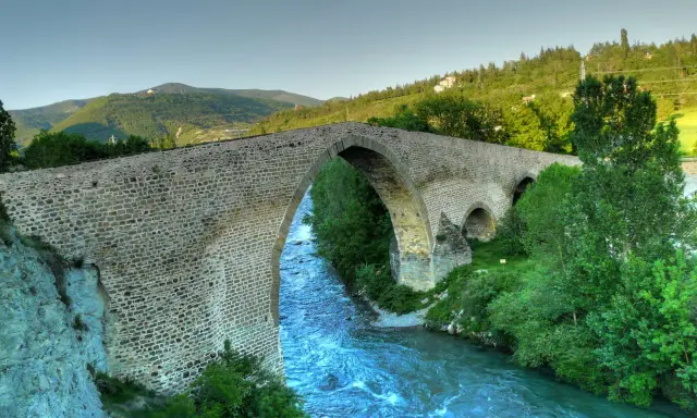 El histórico puente de San Miguel en Jaca cruza un río caudaloso rodeado de vegetación y montañas.