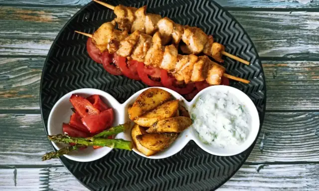 Dwa szaszłyki souvlaki z kurczaka na talerzu z pomidorami, ziemniakami, szparagami i sosem.
