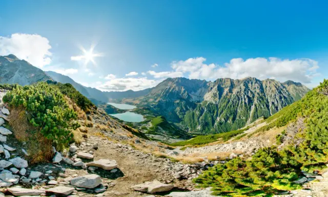 Słoneczna panorama Tatr z dwoma jeziorami w dolinach. Kamienista ścieżka prowadzi przez zielone zbocza porośnięte kosodrzewiną.