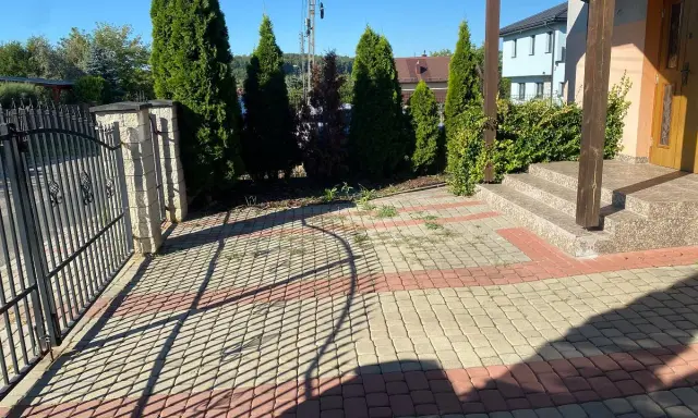 Nowy pomysł na stary chodnik: kostka brukowa w odcieniach beżu i czerwieni tworzy ciekawy wzór przed domem, otoczonym zielenią i ogrodzeniem.