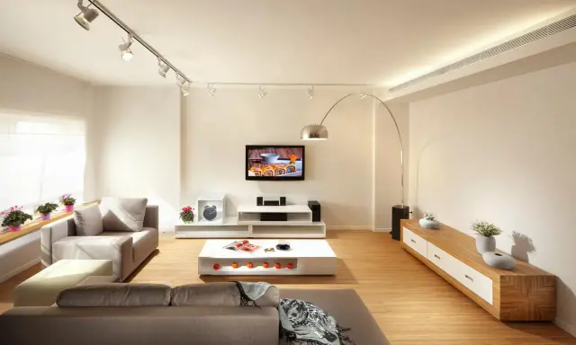 Un salón moderno con sofás grises, mesa de centro blanca y TV. La iluminación, con focos en riel y lámpara de arco, es clave para como iluminar un salón.
