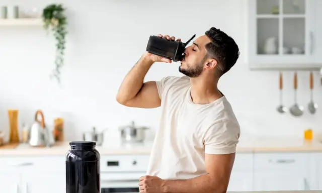 Mann trinkt Proteinshake aus schwarzem Shaker. Daneben steht ein großer Behälter mit Pulver. Wozu Protein**pulver**? Für Muskelaufbau und Erholung.