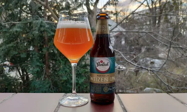 Szklanka z bursztynowym piwem Grolsch Royale Weizen IPA i butelka obok, na tle drzew i nieba.
