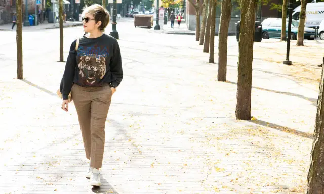 A woman walks down a tree-lined street in a trendy plaid pants outfit, featuring a graphic sweatshirt and white sneakers.