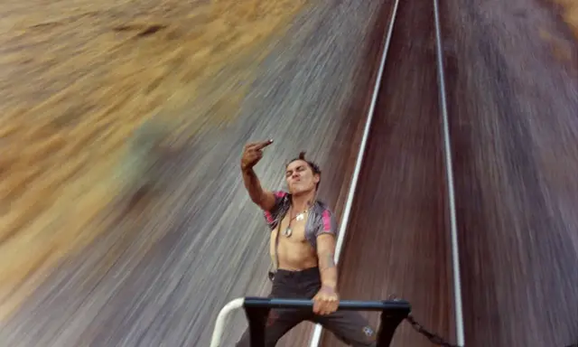 Mike Brodie, un giovane ribelle, fa il gesto del dito medio mentre viaggia su un treno merci, sfidando il mondo con uno sguardo provocatorio.