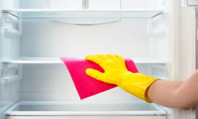 Mano con guante amarillo limpiando estante de frigorífico vacío con paño rosa.