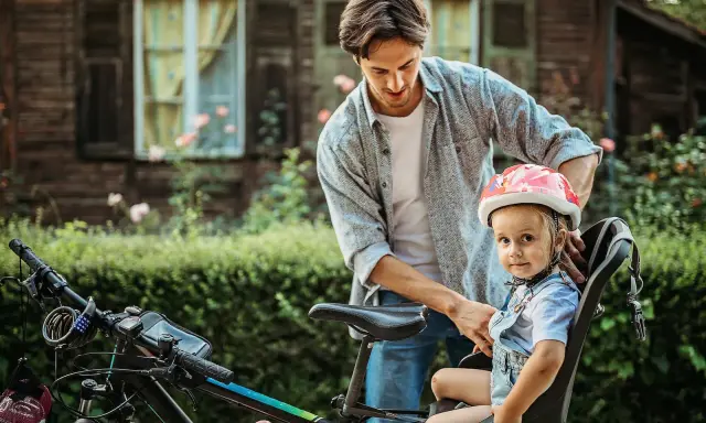 Tata zapina kask dziewczynce, która siedzi w foteliku rowerowym. Od kiedy można wozić dziecko na rowerze? Od kiedy jest bezpiecznie i ma kask!