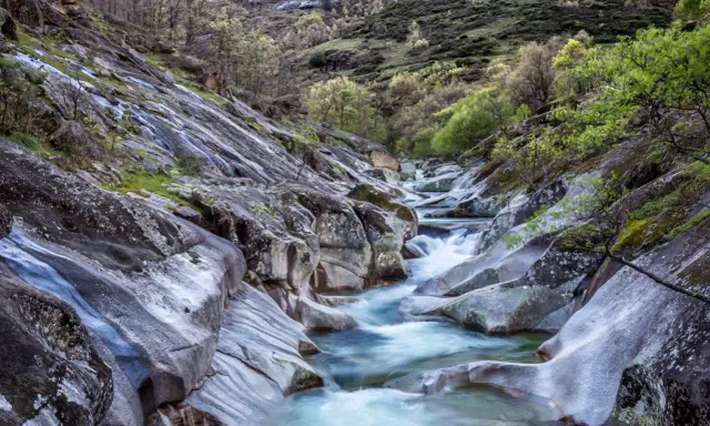Un río de aguas cristalinas serpentea entre rocas pulidas y vegetación exuberante. Un paisaje idílico que evoca la frescura del verano en Extremadura.