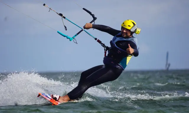 Uśmiechnięty mężczyzna w żółtym kasku i piance cieszy się jazdą na desce. To świetna nauka kitesurfingu!