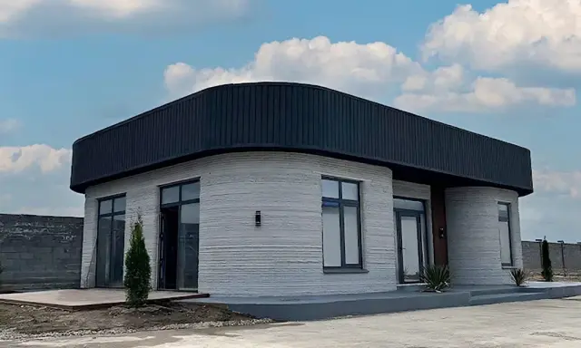Ein modernes 3d druck haus mit runden Ecken und einem dunklen Dach steht unter einem blauen Himmel mit Wolken.