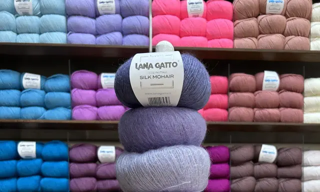 En stapel med tre härvor av lyxigt mohair silke garn i olika lila nyanser. Bakom syns hyllor fyllda med garn i många färger.