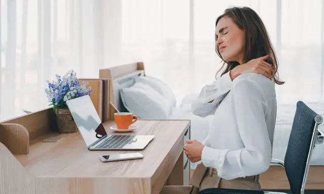 Femme se massant l'épaule, assise à son bureau. La mauvaise position bureau peut causer des douleurs.