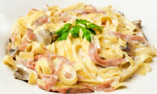 Tagliatelle z szynką i pieczarkami w kremowym sosie, z dodatkiem mozzarelli i natki pietruszki.