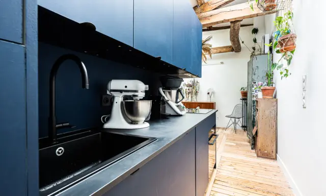 Cuisine moderne avec armoires bleues, évier noir et robot pâtissier. L'entrée de la maison donne directement dans cette pièce fonctionnelle et stylée.