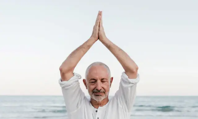 Mężczyzna z zamkniętymi oczami, ręce złożone nad głową, praktykuje mindfulness co to. Spokój nad morzem.