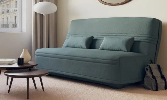 Canapé bz ou clic clac bleu canard, avec coussins, près d'une fenêtre et de tables basses. Parfait pour un petit espace.