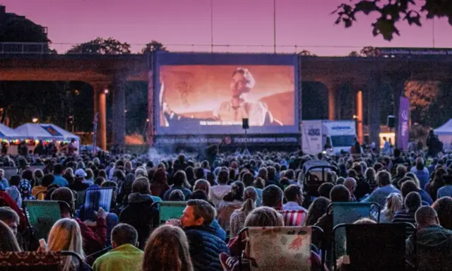 Stor publik njuter av utomhusbio i Stockholm under en rosa kvällshimmel. En stor duk visar en filmscen.