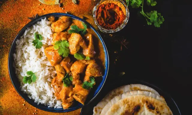 Aksamitny kurczak w sosie butter masala, podany z ryżem i chlebkami naan. Obok miseczka z przyprawami.