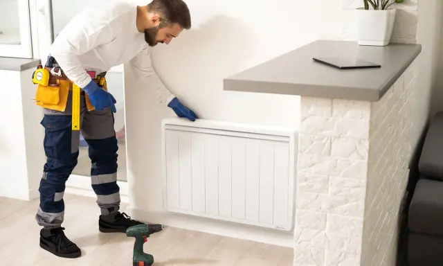 Un technicien en bleu, avec une ceinture à outils, installe un radiateur à inertie. Il est prêt à brancher le radiateur inertie sur prise de courant.