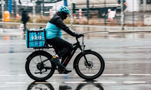Kurier na rowerze z niebieską torbą Wolt, jadący w deszczu. To przesyłka bez możliwości śledzenia, dostarczana szybko i sprawnie.