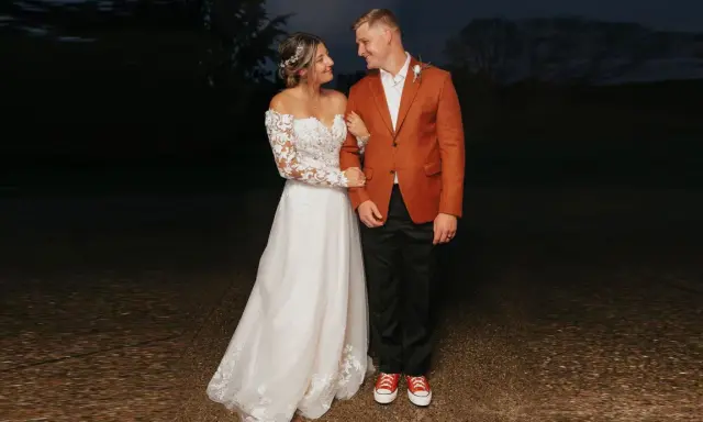 A bride in a lace gown and groom in a rust-colored jacket and black pants, sporting red sneakers, showcase their semi-formal attire for wedding.