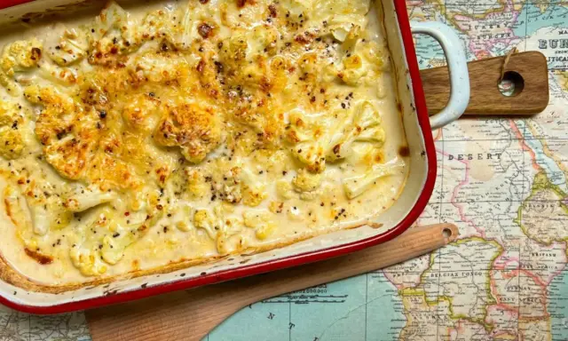 Coliflor gratinado con bechamel cremosa, cubierto de queso dorado y pimienta negra, servido en una fuente roja sobre un mapa antiguo.