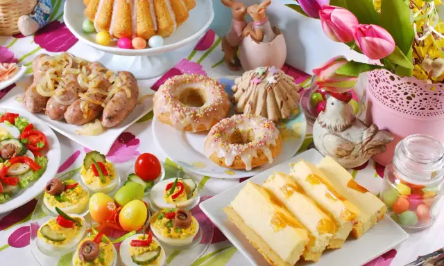 Kolorowy stół z pysznym śniadaniem wielkanocnym: babka, kiełbaski, sałatka, babeczki, sernik i jajka faszerowane.