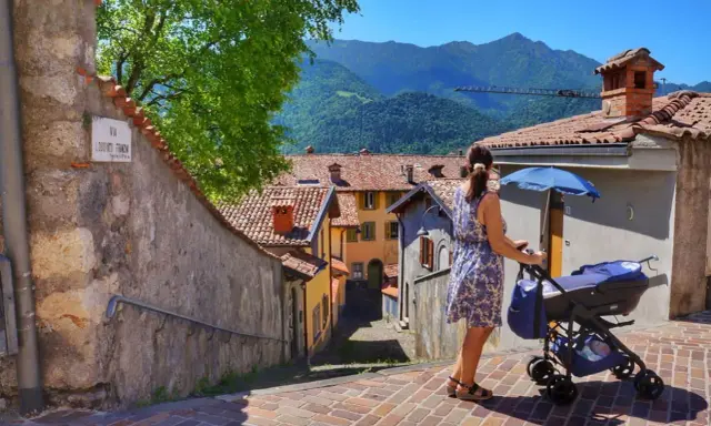 Una donna spinge un passeggino con neonati in montagna, tra case con tetti rossi e un panorama mozzafiato.