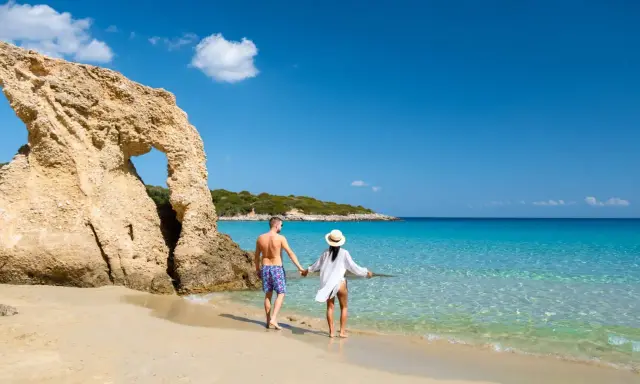 Para, trzymając się za ręce, spaceruje po piaszczystej plaży z turkusową wodą. Czy Cypr jest bezpieczny? Tak, to idealne miejsce na wakacje.