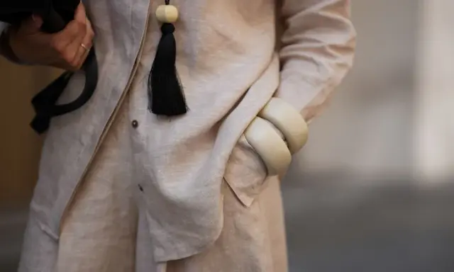 Dettaglio di un outfit in lino materiale, con una mano che tiene una borsa nera, un ciondolo con nappa nera e bracciali color crema.