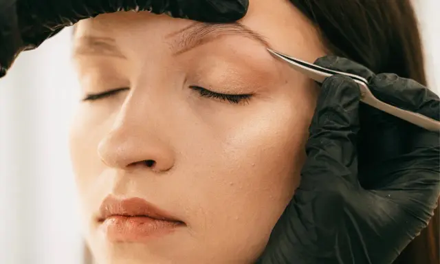 Une main gantée retire délicatement un poil incarné au sourcil d'une femme aux yeux fermés.