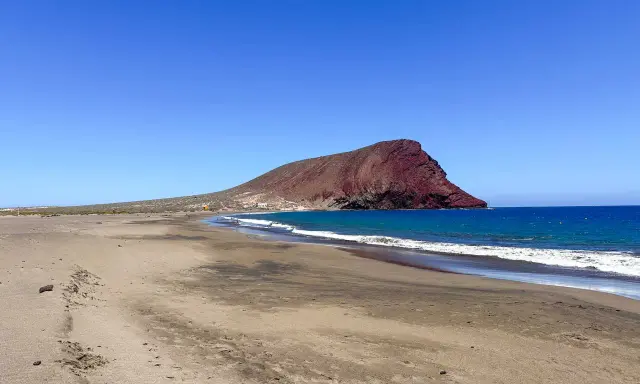 Plaże z białym piaskiem na Teneryfie, z charakterystyczną czerwoną górą w tle i turkusowym oceanem.