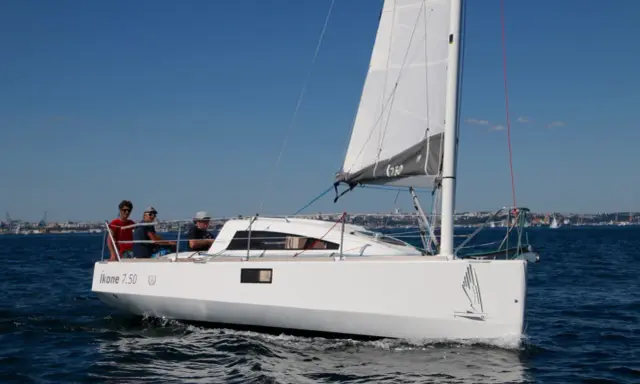 Un voilier Ikone 7.50 navigue par beau temps, avec trois personnes à bord. La voile est hissée, le ciel est bleu.