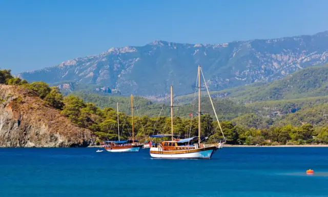 Dwa drewniane jachty kołyszą się na turkusowych wodach, zalesione wzgórza i góry w tle. Idealny dzień na zwiedzanie wybrzeża w okolicach **Antalya miasto**.