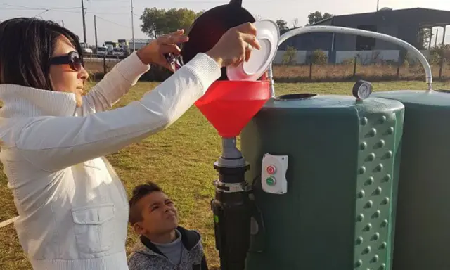 Une femme verse des déchets organiques dans un entonnoir rouge, alimentant un méthaniseur domestique. Un garçon regarde, curieux.