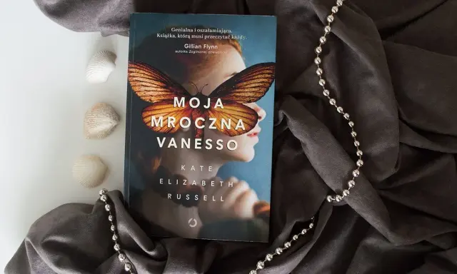 Okładka książki "Moja mroczna Vanesso" z motywem motyla na twarzy dziewczyny, otoczona szarym materiałem i koralikami.